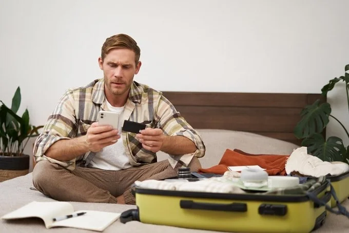 Man packing a suitcase and planning a slow travel trip while sitting on a bed with thoughtful preparation.