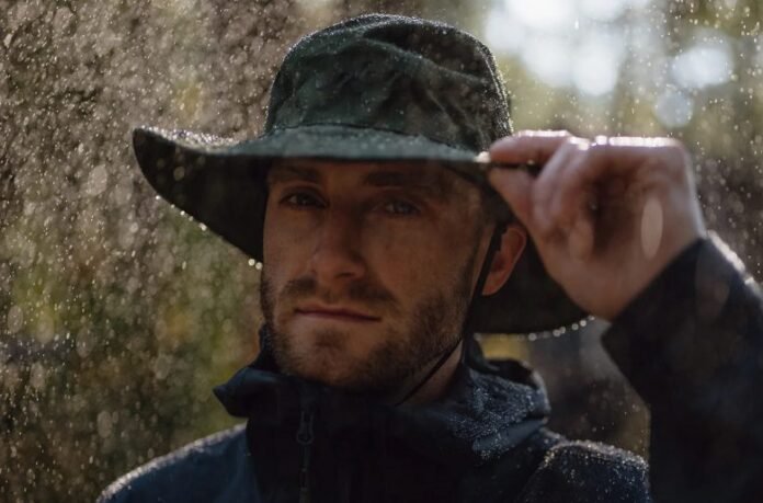 Man adjusting wide-brimmed dark green rain hat while water droplets fall