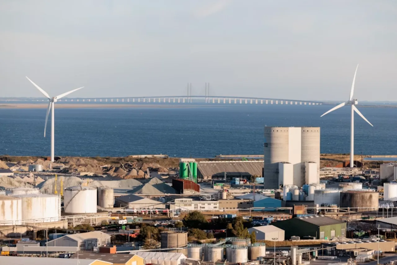 Coastal industrial facility with wind turbines, fuel storage tanks, and a long sea bridge in the background promoting renewable energy infrastructure.